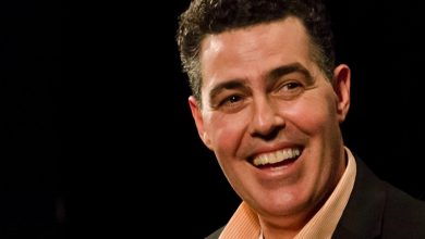 Adam Carolla wearing a suit and tie smiling and looking at the camera