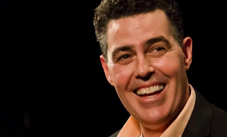 Adam Carolla wearing a suit and tie smiling and looking at the camera